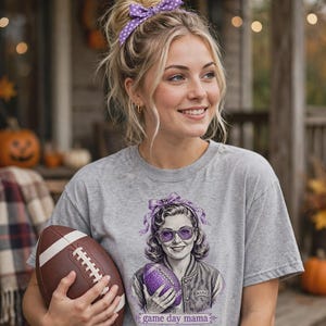 Peut inclure: Un t-shirt gris chiné à manches courtes avec une impression graphique représentant une personne portant un nœud et des lunettes violets, tenant un ballon de football violet. Le texte game day mama est inscrit en dessous.