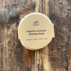 May include: A round, white bar of organic lavender shaving soap wrapped in brown paper with the text "The Handmade Haven" and "Organic Lavender Shaving Soap" printed on it. The text "Handmade - Organic - Natural - Vegan" is also printed on the paper.