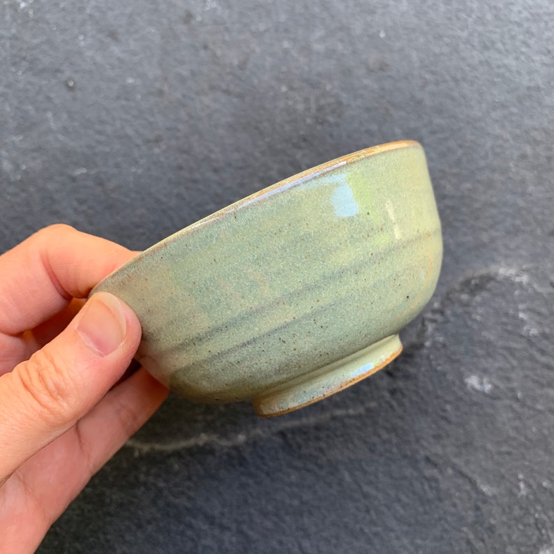 May include: A small, light green ceramic bowl with a slightly uneven glaze. The bowl has a rounded shape and a slightly flared rim.