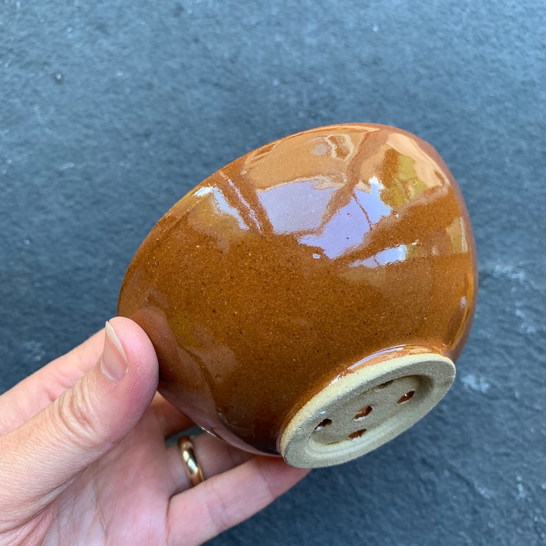 May include: A brown ceramic bowl with a glazed finish and a drainage hole at the bottom.