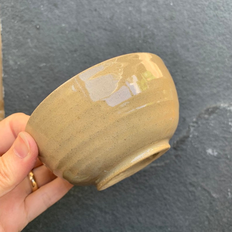 May include: A small, beige ceramic bowl with a slightly textured surface. The bowl has a rounded shape and a slightly flared rim.