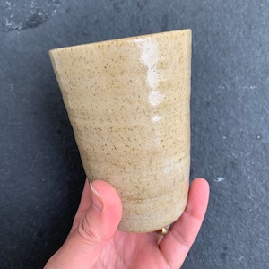 May include: A beige ceramic mug with a speckled glaze. The mug has a rounded shape and a slightly flared lip.