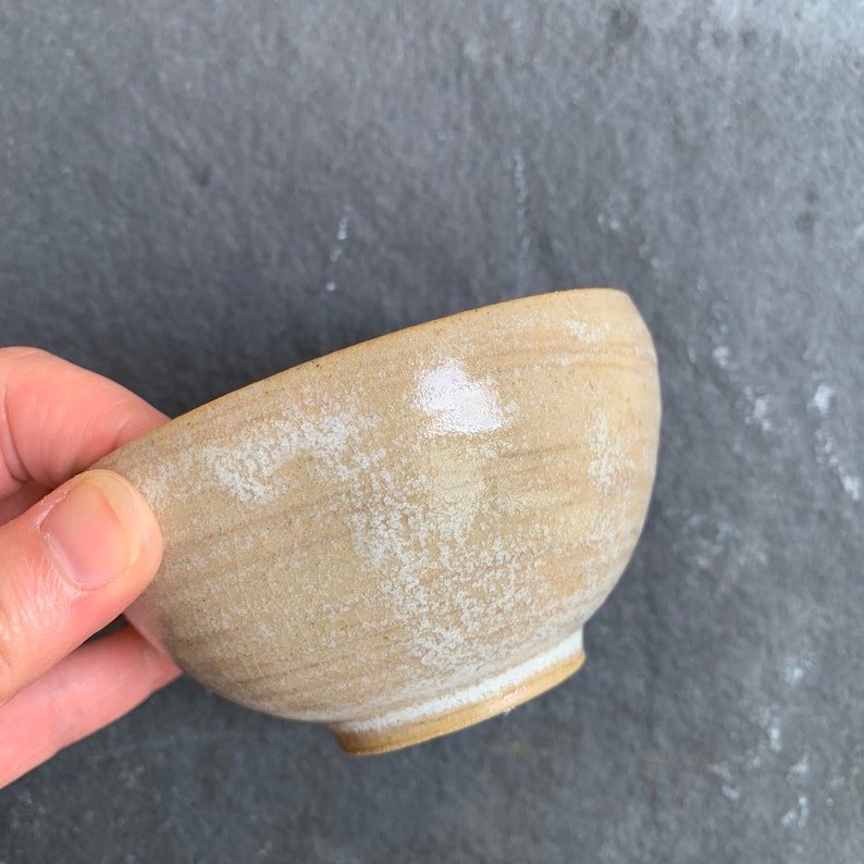 May include: A small, beige ceramic bowl with a white glaze. The bowl has a slightly irregular shape and a textured surface.