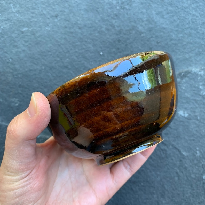 May include: A dark brown ceramic bowl with a glossy finish. The bowl has a rounded shape and a slightly flared rim.