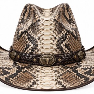 Könnte beinhalten: Ein Cowboyhut mit einem beigen und dunkelbraunen Schlangenledermuster. Der Hut verfügt über ein dunkelbraunes, geflochtenes Lederband, das mit Metallnieten und einem ovalen Messingemblem in Antik-Optik mit einem Stierschädel verziert ist.