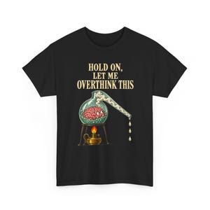 May include: A black t-shirt featuring the text "HOLD ON, LET ME OVERTHINK THIS" in cream letters above a graphic of a pink brain inside a glass laboratory flask. The flask sits on a stand over a small gold oil lamp with a bright orange flame.