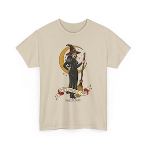 May include: A beige short-sleeve t-shirt featuring a graphic of a witch in a black dress and hat holding a broomstick in front of a yellow crescent moon. A red banner below reads "THEY MISSED ONE" with the text "SALEM 1692" printed underneath.