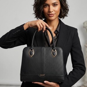 Könnte beinhalten: Eine strukturierte schwarze Handtasche mit texturierter Oberfläche, ausgestattet mit zwei abgerundeten Tragegriffen, die an ovalen goldfarbenen Beschlägen befestigt sind. Vorne steht das Wort Cara in kleinen goldenen Buchstaben.
