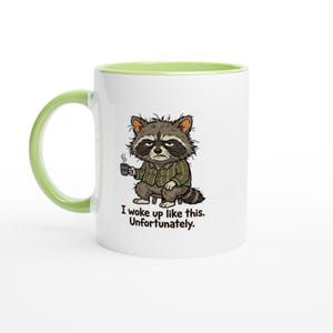 Könnte beinhalten: Ein weißer Keramikbecher mit hellgrünem Henkel und Innenrand. Der Becher zeigt eine Cartoon-Illustration eines zerzausten Waschbären in einem gestreiften Hemd, der eine kleine Tasse hält, mit dem Text: I woke up like this. Unfortunately.