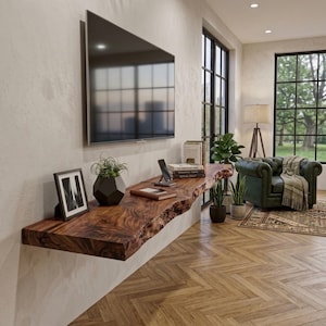 Op de afbeelding: Een lange, aan de muur gemonteerde zwevende plank van natuurlijk hout met een boomkant, voorzien van een donkerbruin nerfpatroon. De plank bevat een zwarte fotolijst, een geometrische zwarte plantenbak met een groene plant, een smartphone en boeken.