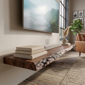 Könnte beinhalten: Ein schwebendes Wandregal aus dunkelbraunem Holz mit natürlicher Baumkante, auf dem ein Stapel aus drei Büchern mit den Titeln Interior Design, Sustainable Living und Woodworking Craft unter einem wandmontierten Fernsehbildschirm liegt.