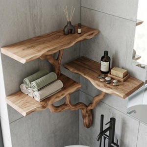 May include: Two tiered wall mounted shelves made of natural wood with live edges and organic branch shaped supports. The shelves hold rolled sage green and cream towels, a dark brown diffuser, a glass bottle, a bar of soap, and small smooth stones.