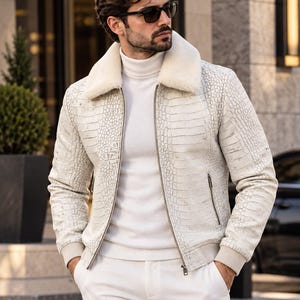 Könnte beinhalten: Eine weiße Jacke mit texturiertem Krokodilleder-Muster, ausgestattet mit einem cremefarbenen Kunstfellkragen, einem durchgehenden Frontreißverschluss und gerippten Bündchen. Die Jacke wird über einem weißen Rollkragenpullover und einer weißen Hose getragen.