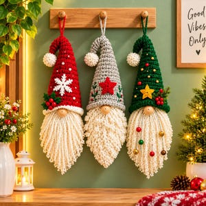 Puede incluir: Tres adornos de gnomos de ganchillo colgantes con largas barbas blancas. El gnomo de la izquierda tiene un gorro rojo con un copo de nieve blanco, el del medio un gorro gris con una estrella roja y el de la derecha un gorro verde con una estrella amarilla.