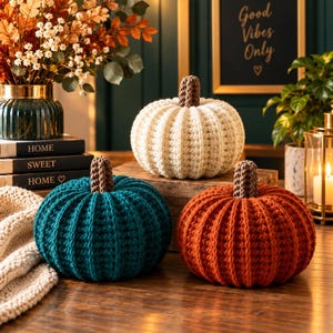 May include: Three round, crocheted pumpkins in cream, teal, and burnt orange, each featuring a textured, ribbed surface and a brown, braided stem. These soft, handcrafted decorative items are arranged on a wooden surface, showcasing distinct colors.