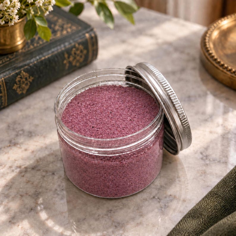 Open jar of Raspberry Bloom pink quartz sand with its silver lid leaning against it, on a marble surface next to an old green book, a small brass vase with white flowers and a brass tray