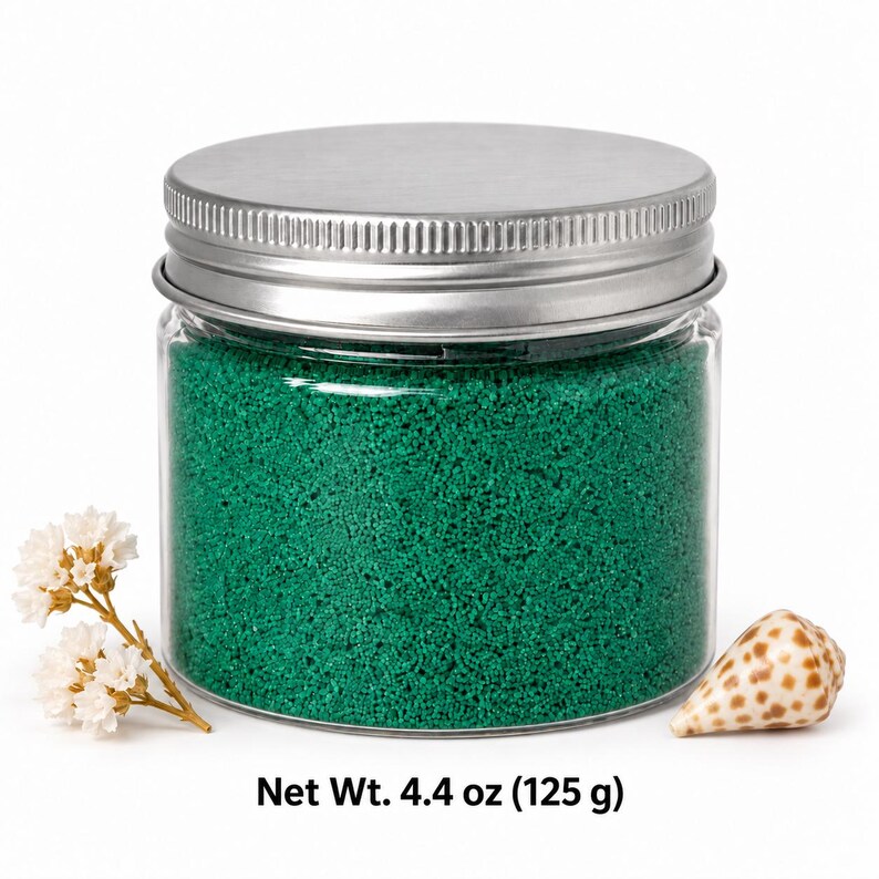 Closed clear jar of Lagoon Green colored quartz sand in a rich emerald green, with a silver metal screw lid, next to small dried white flowers and a spotted cone seashell, with text: Net Wt. 4.4 oz (125 g).