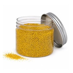 Open clear jar of mustard yellow colored quartz sand with its silver metal screw lid leaning behind it and a small pile of the same sand spilled in front, showing the fine grain texture, on a white background.