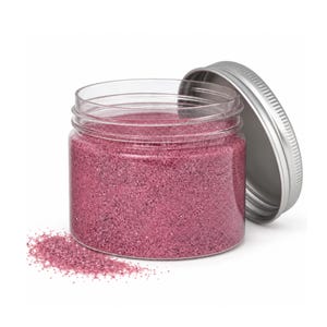 Open clear jar of soft raspberry pink colored quartz sand with its silver metal screw lid leaning behind it and a small pile of the same sand spilled in front, showing the fine grain texture, on a white background.