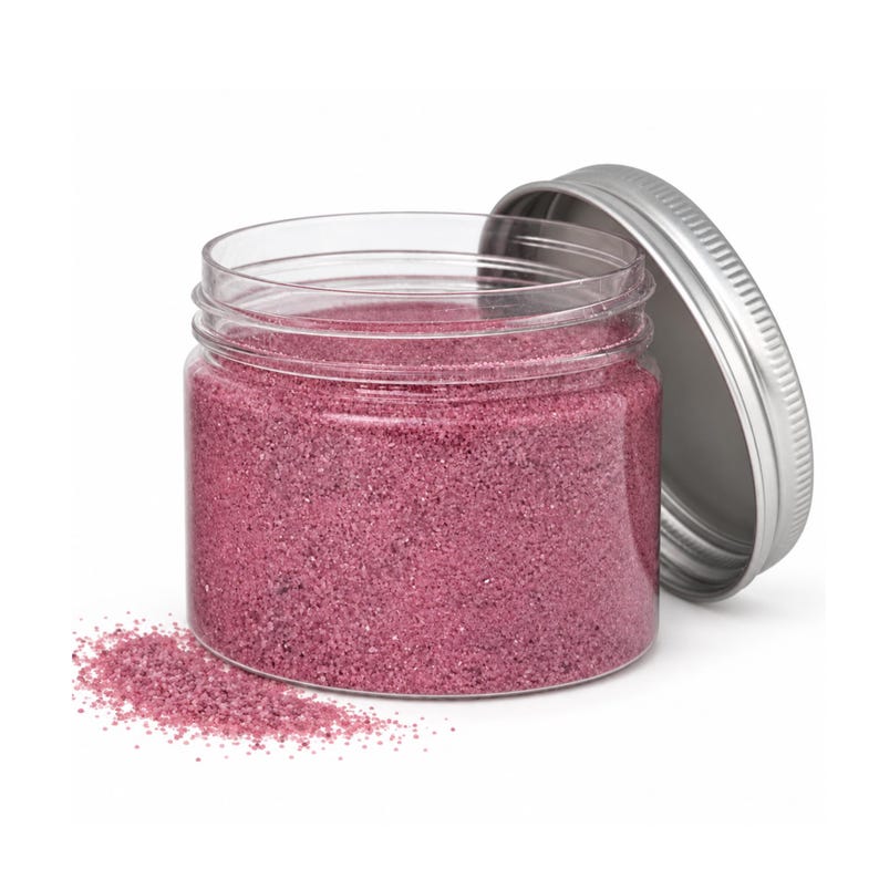 Open clear jar of soft raspberry pink colored quartz sand with its silver metal screw lid leaning behind it and a small pile of the same sand spilled in front, showing the fine grain texture, on a white background.