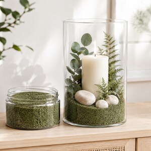 Tall glass cylinder holder with a lit white pillar candle, eucalyptus, fern and white pebbles set in Olive Grove green quartz sand, next to an open jar of the same sand on a light wooden cabinet