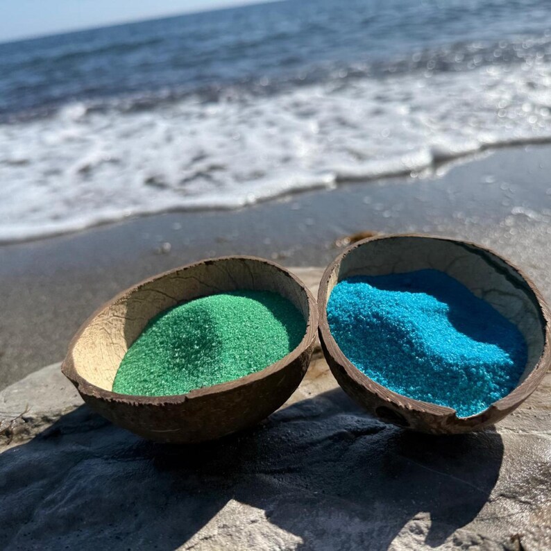 Two coconut shell bowls on a beach, one with green craft sand and one with bright blue sand