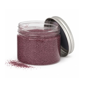 Open clear jar of dusty plum mauve colored quartz sand with its silver metal screw lid leaning behind it and a small pile of the same sand spilled in front, showing the fine grain texture, on a white background.