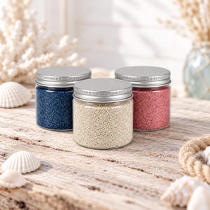 Op de afbeelding: Drie doorzichtige cilindrische potten met zilveren schroefdeksels, elk gevuld met fijnkorrelig zand. De linkerpot bevat donkerblauw zand, de middelste pot beige zand en de rechterpot roze zand.