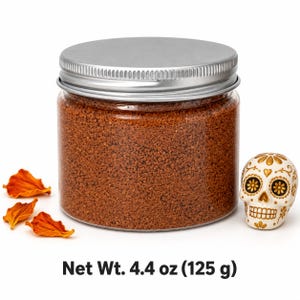 Closed clear jar of Terracotta Clay colored quartz sand in a warm reddish brown, with a silver metal screw lid, next to dried orange flower petals and a white and gold decorated sugar skull figurine, with text: Net Wt. 4.4 oz (125 g).