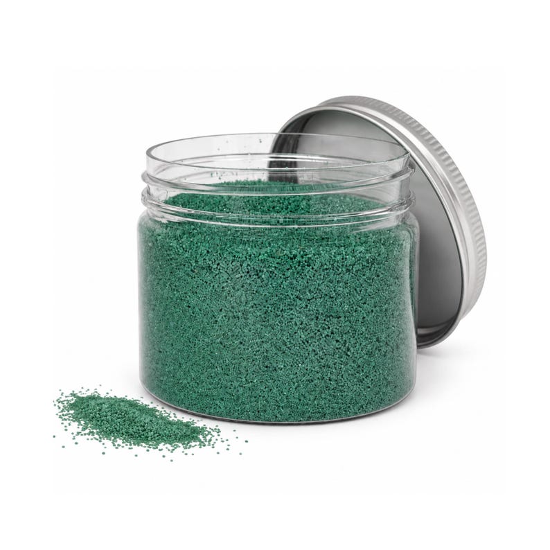 Open clear jar of emerald green colored quartz sand with its silver metal screw lid leaning behind it and a small pile of the same sand spilled in front, showing the fine grain texture, on a white background.