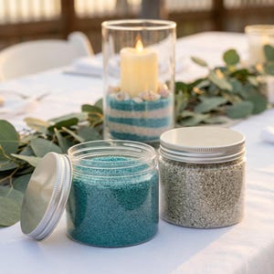 Open jar of teal craft sand and a jar of gray sand on a white table, with a candle in a teal and gray striped sand holder
