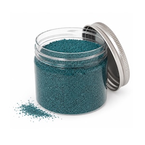 Open jar of dark teal craft sand with its silver lid leaning on the side and a little sand spilled in front, on a white background