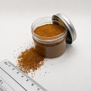 Open jar of Terracotta Clay orange brown quartz sand with its silver lid, next to a small pile of spilled sand and a ruler in centimeters showing the fine grain size.