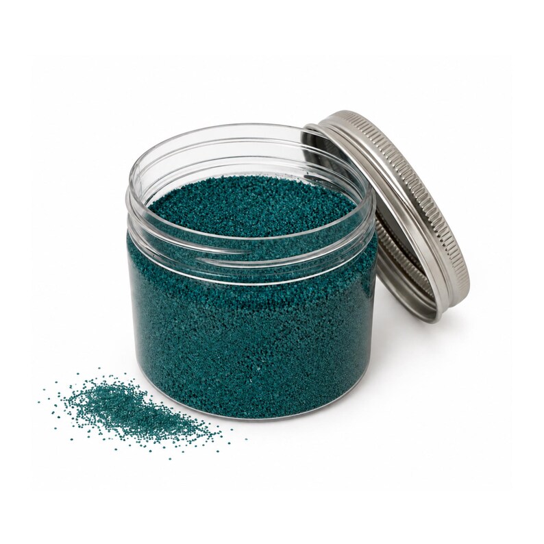 Open clear jar of deep teal colored quartz sand with its silver metal screw lid leaning behind it and a small pile of the same sand spilled in front, showing the fine grain texture, on a white background.
