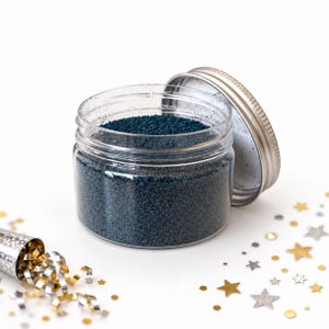 Open clear jar of deep blue colored quartz sand with the silver metal lid leaning against it, next to a silver party popper spilling gold and silver curly ribbons, and scattered gold and silver star confetti, on a white background.