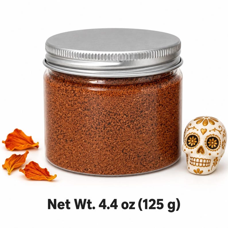 Closed clear jar of terracotta brown colored quartz sand with a silver metal screw lid, next to dried orange flower petals and a white and gold decorated sugar skull figurine, with text: Net Wt. 4.4 oz (125 g).
