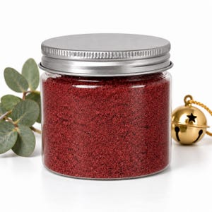 Closed clear jar of deep red colored quartz sand with a silver metal screw lid, next to a eucalyptus sprig and a gold jingle bell on a gold cord, on a white background.