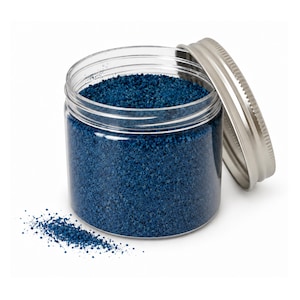 Open jar of deep blue craft sand with its silver lid leaning on the side and a small pile of sand spilled in front, on a white background