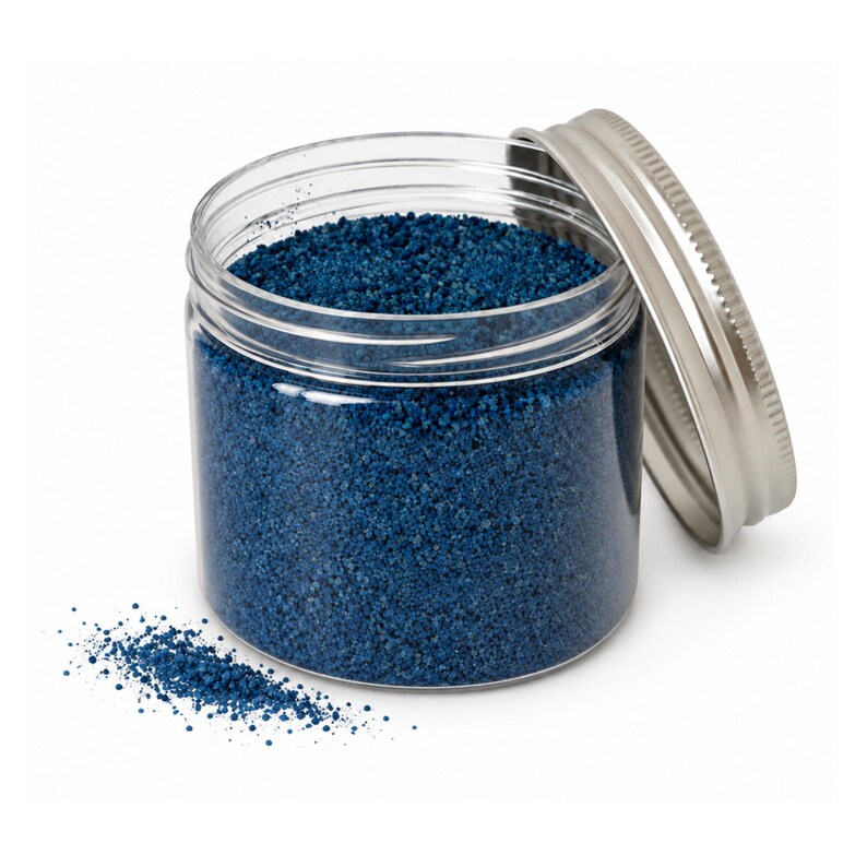 Open jar of deep blue craft sand with its silver lid leaning on the side and a small pile of sand spilled in front, on a white background
