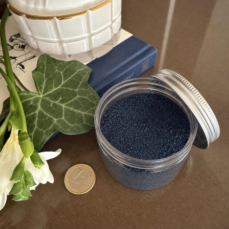 Open jar of Atlantic Night craft sand next to a 1 euro coin for size, with white flowers, ivy leaf, and a blue book