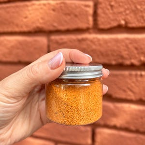 K&ouml;nnte beinhalten: Ein kleiner, zylindrischer Beh&auml;lter aus klarem Kunststoff, gef&uuml;llt mit einer leuchtend orangefarbenen, k&ouml;rnigen Gew&uuml;rzmischung. Der Beh&auml;lter ist mit einem einfachen, runden, silberfarbenen Schraubdeckel aus Metall mit geriffeltem Rand versehen.