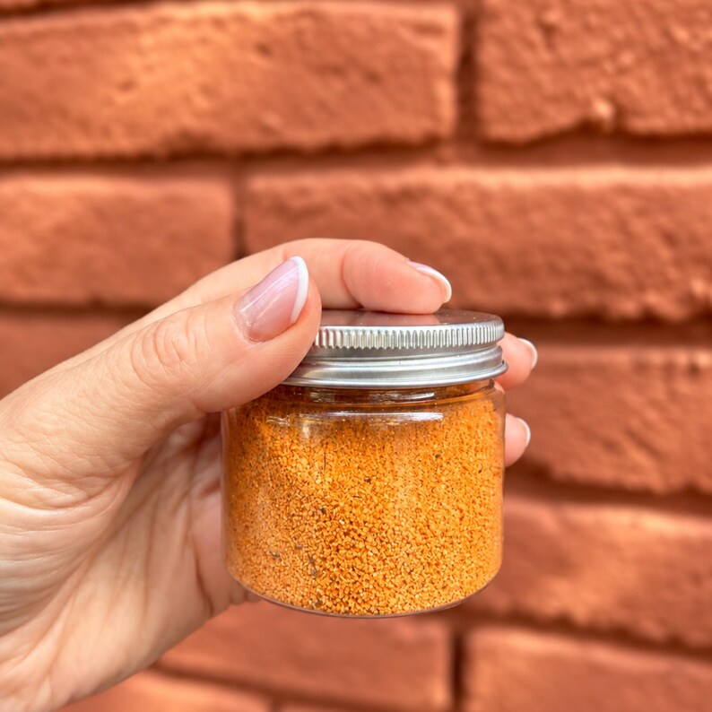K&ouml;nnte beinhalten: Ein kleiner, zylindrischer Beh&auml;lter aus klarem Kunststoff, gef&uuml;llt mit einer leuchtend orangefarbenen, k&ouml;rnigen Gew&uuml;rzmischung. Der Beh&auml;lter ist mit einem einfachen, runden, silberfarbenen Schraubdeckel aus Metall mit geriffeltem Rand versehen.