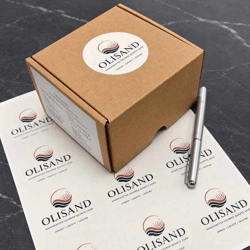 Peut inclure: Une bo&icirc;te en carton brun avec un autocollant circulaire portant le logo et le texte Olisand. Une feuille d'autocollants circulaires assortis se trouve sous la bo&icirc;te, et un stylo en m&eacute;tal argent&eacute; repose sur la feuille &agrave; c&ocirc;t&eacute; de la bo&icirc;te.