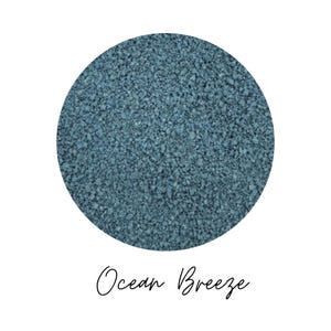 Close-up circle of Ocean Breeze colored quartz sand showing fine teal blue grains, with the color name Ocean Breeze written in script below, on a white background