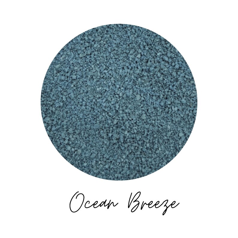 Close-up circle of Ocean Breeze colored quartz sand showing fine teal blue grains, with the color name Ocean Breeze written in script below, on a white background