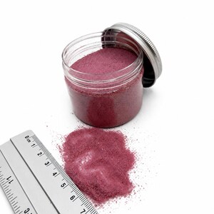 Open jar of Raspberry Bloom pink quartz sand with its silver lid, next to a small pile of spilled sand and a ruler in centimeters showing the fine grain size.
