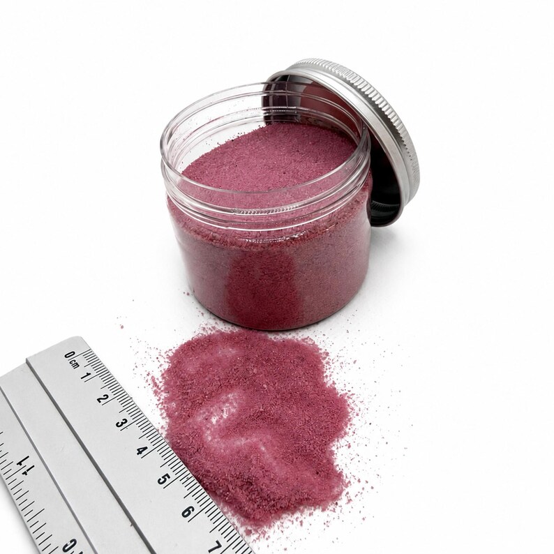 Open jar of Raspberry Bloom pink quartz sand with its silver lid, next to a small pile of spilled sand and a ruler in centimeters showing the fine grain size.