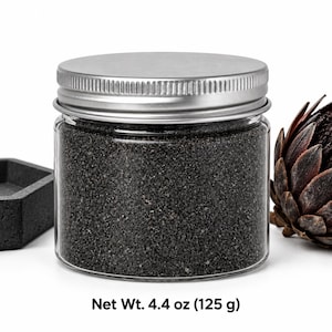 Closed clear jar of charcoal black colored quartz sand with a silver metal screw lid, next to a dark concrete tray and a dried dark flower head, with text: Net Wt. 4.4 oz (125 g).