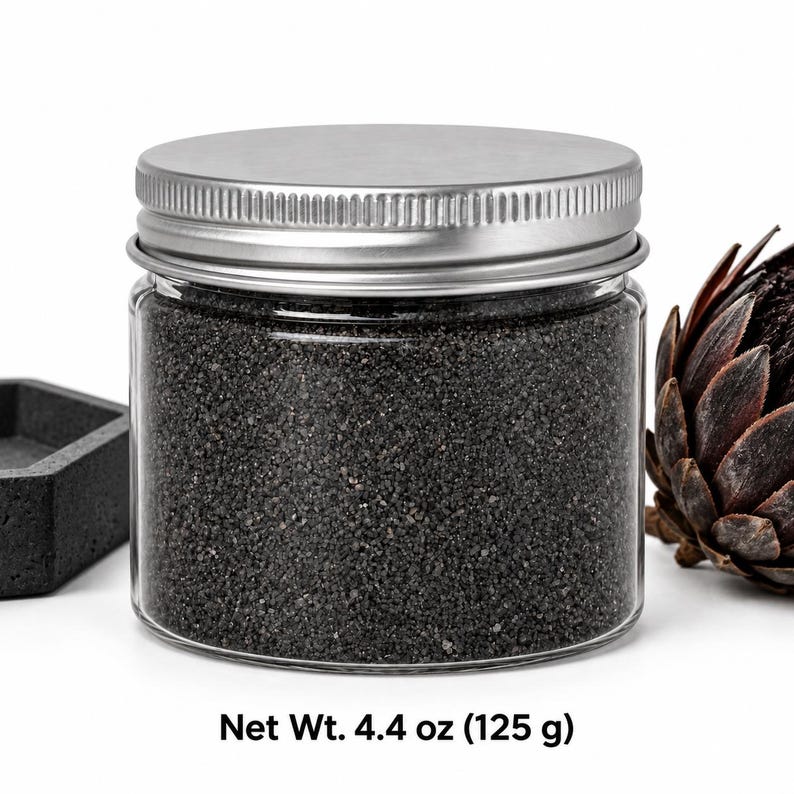 Closed clear jar of charcoal black colored quartz sand with a silver metal screw lid, next to a dark concrete tray and a dried dark flower head, with text: Net Wt. 4.4 oz (125 g).