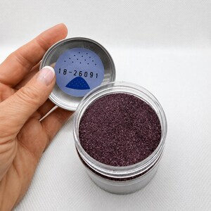 Top view of an open jar of Plum Velvet plum quartz sand, with a hand holding the silver lid, which has a label with the number 18-26091 and a small sand pile illustration on the inside